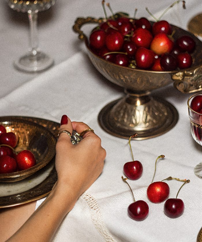 A beautiful table is adorned with silver bowls filled with ripe cherries while a hand picks one. The atmosphere is warm and inviting, creating a sense of classic elegance.